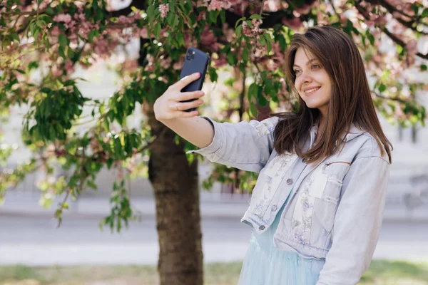 Fiatal mosolygós nő, aki önarcképeket készít az okostelefonján. Modell pózol park sakura fák háttér. Nő pozitív arc érzelmek. — Stock Fotó