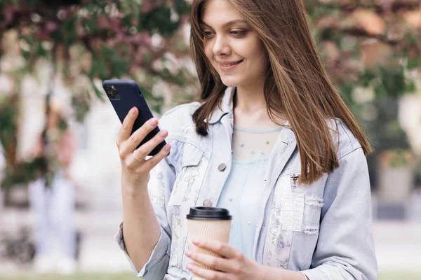Atrakcyjna młoda kobieta ze smartfonem i filiżanką kawy w rękach na tle drzewa sakura w parku miejskim. Ładna, letnia kobieta w białej sukience patrzy na telefon i uśmiecha się. Stylowy strój — Zdjęcie stockowe
