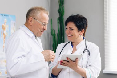 Tıbbi ekip iki yaşlı erkek ve genç bayan doktor beyaz önlüklü konuşmalar giyer, dijital tablet kullanır. Tıp konseptinde iki hastane ortaklığı, sağlık teknolojisi.