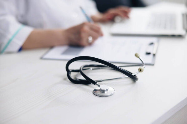 Close up of stethoscope medical equipment with female doctor working in background. Professional physician wearing white coat writing notes using laptop at desk