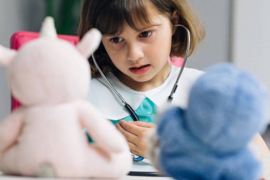 Küçük sevimli kreş çocuğu elinde steteskop tutarak hasta bir oyuncak hastayı dinlerken sağlık üniforması giyiyor. Küçük akıllı çocuk hastane oyunu oynuyor. Doktor rolü yapıyor..