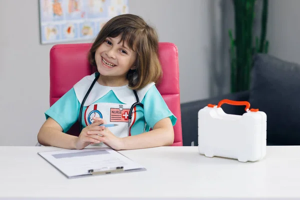 Retrato de enfermera médica con uniforme médico mirando a la cámara y sonriendo. Linda adorable niña preescolar pequeña sonriendo en el hospital —  Fotos de Stock