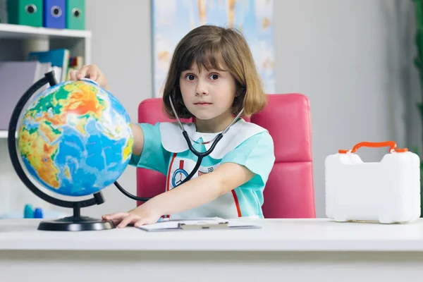 Little girl is dressed in medical clothes with a stethoscope in her hands examines and plays a game of healing the planet Earth. World planet earth eco friendly. Sick earth planet Covid-19 symptoms.