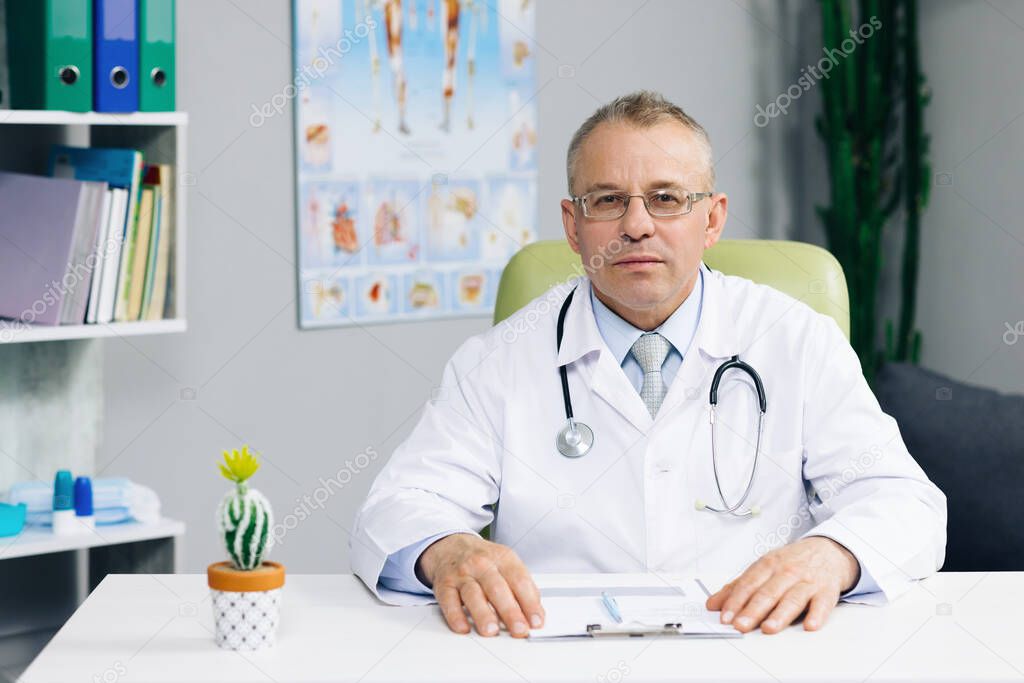 Portrait of confident old mature male head doctor physician in white medical uniform in glasses sitting at workplace. General practitioner looking at camera, posing in office