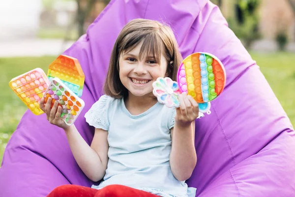 Menina retrato com um brinquedo popit moderno. Colorido e brilhante pop-lo brinquedo. Simples covinha. A criança está sentada em uma cadeira fácil com brinquedo sensorial antiestresse colorido da moda pop-lo e covinha simples — Fotografia de Stock