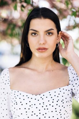 Sokak şehri yaz kızı şehrinde kameraya bakan, çekici, güzel saçlı, ciddi genç bir kadınla yüzleş. Şehir parkında bahar çiçekleri.