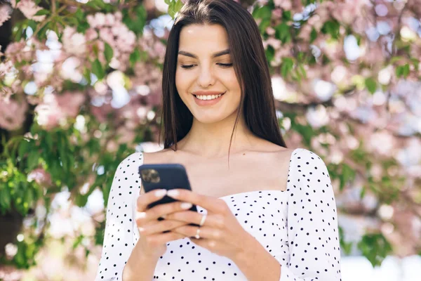 Uśmiechnięta kobieta idzie ulicą Central Parku i używa telefonu. Pretty Summer kobieta w białej sukience idzie ulicą patrząc na jej telefon komórkowy — Zdjęcie stockowe