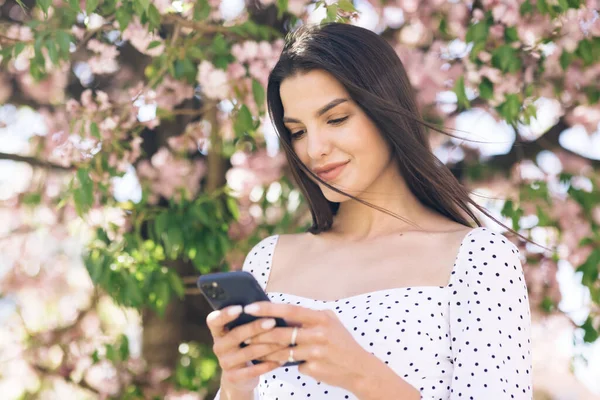 Portret pięknej kobiety korzystającej ze smartfona w parku na tle drzew sakura. Młoda piękna dziewczyna przeglądająca Internet za pomocą portali społecznościowych — Zdjęcie stockowe