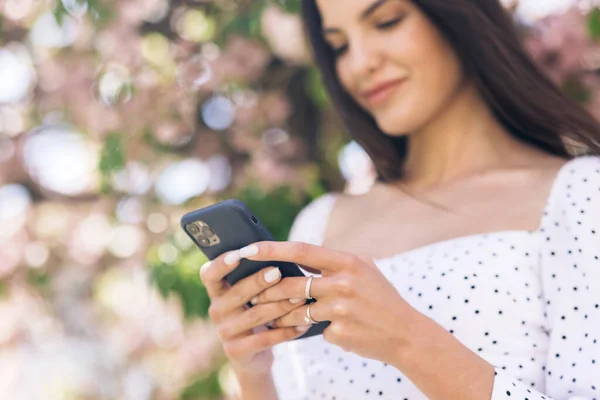 Portret szczęśliwej hipsterki wpisującej przez telefon komórkowy na zewnątrz. Wesoła dziewczyna spacerująca ze smartfonem w tle miejskim. Uśmiechnięta dama trzymająca komórkę w rękach na zewnątrz — Zdjęcie stockowe