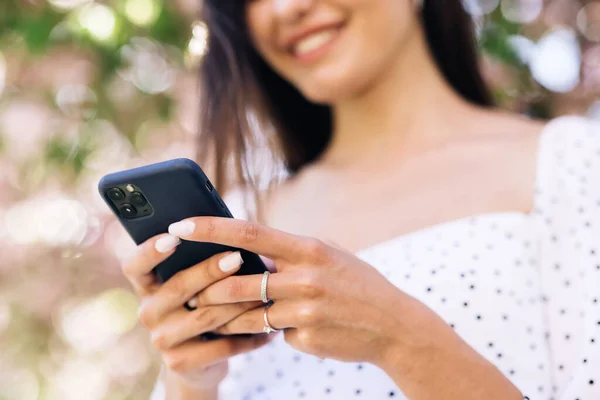 Wesoła dziewczyna ze smartfonem w parku na tle drzew sakura. Uśmiechnięta dama trzymająca komórkę na zewnątrz. Happy hipster kobieta wpisując przez telefon komórkowy na zewnątrz. — Zdjęcie stockowe