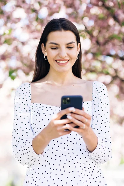 Szczęśliwa hipsterka pisząca przez telefon komórkowy na dworze. Wesoła dziewczyna ze smartfonem w parku na tle drzew sakura. Uśmiechnięta dama trzymająca komórkę w rękach na zewnątrz — Zdjęcie stockowe