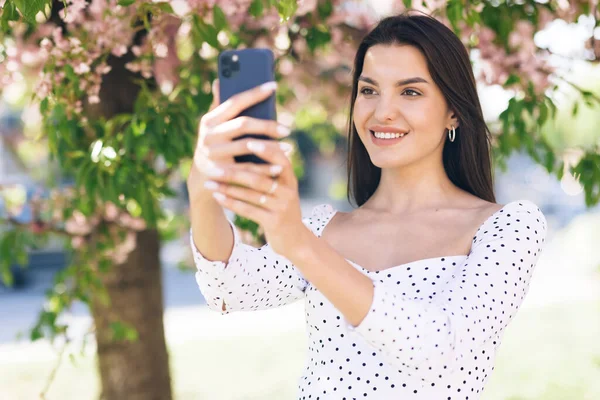 Uśmiechnięta kobieta na smartfonie w mieście, przyjazna kobieta machająca ręką przed kamerą, ładna kobieta na czacie wideo przez telefon, stylowa blogerka nagrywająca wideo przez telefon na zewnątrz — Zdjęcie stockowe