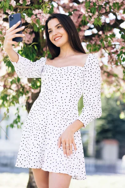 Młoda uśmiechnięta kobieta w letniej białej sukience robiąca selfie zdjęcia autoportretowe na smartfonie. Model pozowanie na tle drzewa sakura parku. Kobieta wykazująca pozytywne emocje na twarzy. — Zdjęcie stockowe