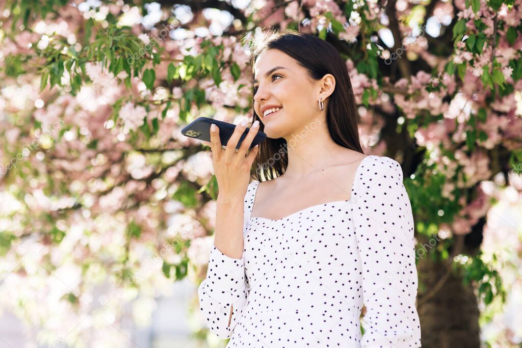 Happy young girl using smart phone voice recognition, dictates thoughts, voice dialing message outdoor talking to mobile assistant. Talking mouth in the speaker of a young woman