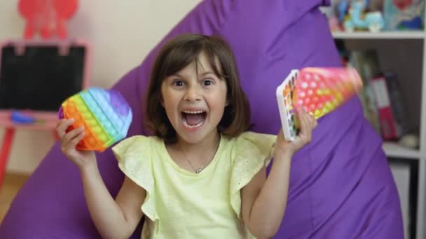 Portret klein meisje met een modern popit speelgoed. Het kind zit op een fauteuil met kleurrijke trendy antistress sensorische speelgoed pop it en eenvoudige kuiltje. Kleurrijk en helder pop het speelgoed. Eenvoudige kuiltje. — Stockvideo