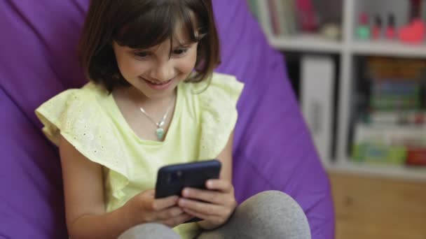 Petite fille caucasienne mignonne tenant téléphone intelligent profiter de l'utilisation d'applications mobiles, jouer à des jeux à la maison. Petit enfant apprenant dans son téléphone portable, regardant la vidéo, s'amusant avec le concept de technologie mobile — Video