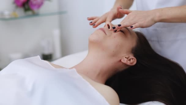 Die junge schöne kaukasische Frau, die auf einem Wellnessbett liegt, erhält eine Gesichtsmassagebehandlung mit ätherischem Aromaöl von einem Masseur im Schönheitssalon. Wellness Gesundheitsfürsorge Körpermassage Spa-Konzept. — Stockvideo