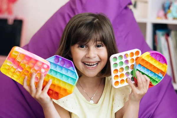 Portrait little girl with a modern popit toy. The child is sitting on a easy chair with colorful trendy antistress sensory toy pop it and simple dimple. Colorful and bright pop it toy. Simple dimple. — Stock Photo, Image