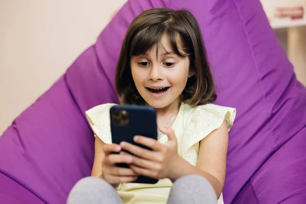 Menina bonito criança segura telefone, criança usando smartphone, criança navegando na Internet, usa a comunicação de vídeo em casa no telefone — Fotografia de Stock