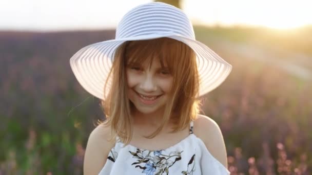 Крупним планом портрет молодої щасливої дівчинки з капелюхом на голові в лавандових полях на фоні. Жінка одягнена в капелюх посміхається навесні — стокове відео
