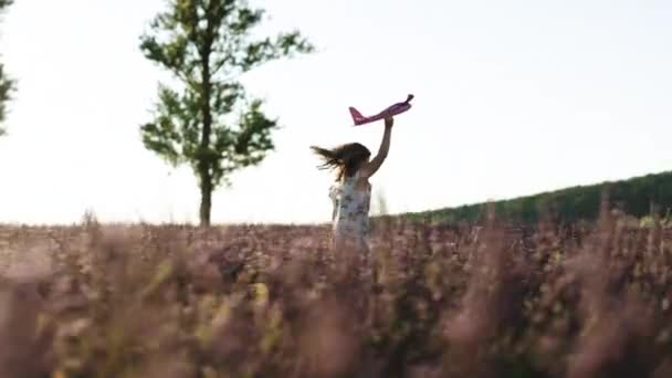 Ragazza con l'aereo nelle mani di correre su una collina al tramonto. Ragazza felice corre con un aeroplano giocattolo su un campo di lavanda alla luce del tramonto. I bambini giocano aereo giocattolo. Concetto grande bambino sogno. — Video Stock