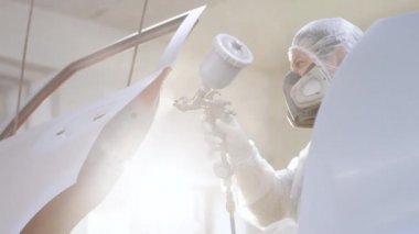 Usta araba servisinde otomobilin bir parçasını boyuyor. Elinde tutarak, koruyucu üniforma giymiş bir şekilde sprey sıkma makinesi kullanıyor. Modern araba boyama.