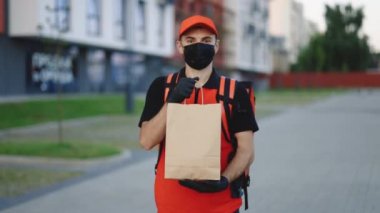 Kırmızı üniformalı, şapkalı ve tıbbi maskeli beyaz bir adamın portresi servis servisinden dönerken kameraya yemek paketi ile gülümsüyor. Çevre marketi paketli erkek kurye.