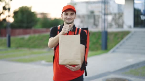 Venkovní portrét doručovatele s červenou uniformou držícího tašky na jídlo čeká na zákazníka. Detailní záběr šťastný mladý kurýr je hrdý na svou práci, usmívá se na ulici. Domácí doručení — Stock video