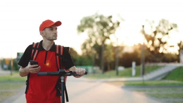 男性信使食物运送与红色热背包步行街电动摩托车使用智能手机导航。送货员员工在日落时提供网上定单客户. — 图库视频影像