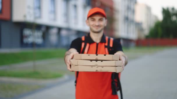 Transportador jovem atraente do homem da entrega no tampão vermelho que sorri na câmera que prende caixas da pizza que entregam o alimento rápido em torno da cidade. Retrato ao ar livre. Trabalhador urbano. Conceito de correio, entrega em domicílio, pizza — Vídeo de Stock