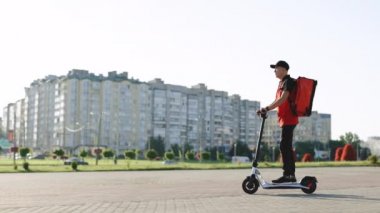 Asyalı kurye yemek teslimatı. Kırmızı termal sırt çantalı. Elektrikli scooter ile sokakta geziyor. Kırmızı üniformalı teslimatçı işçi. Mükemmel atış.