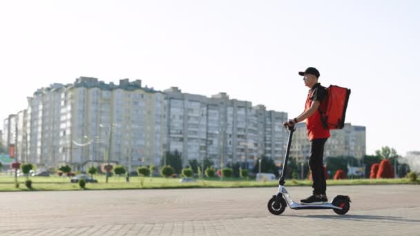 Asyalı kurye yemek teslimatı. Kırmızı termal sırt çantalı. Elektrikli scooter ile sokakta geziyor. Kırmızı üniformalı teslimatçı işçi. Mükemmel atış. — Stok video