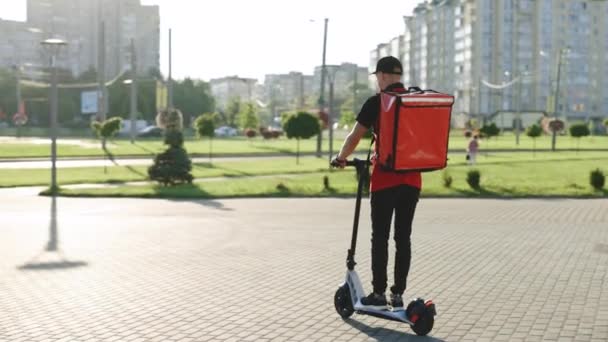 Kırmızı termal sırt çantalı erkek kurye yemek teslimatı elektrikli scooter ile caddeye çıkıp müşteriye sipariş veriyor. Kırmızı üniformalı teslimatçı işçi. Mükemmel atış. — Stok video
