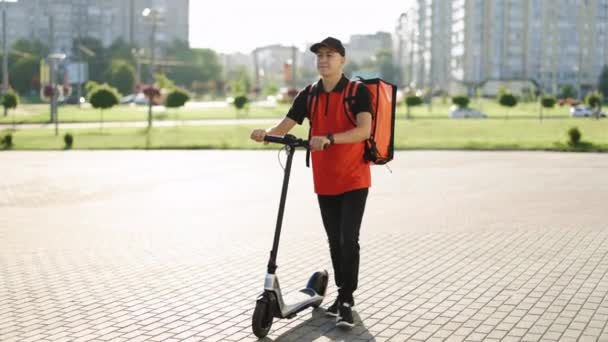 Kırmızı termal sırt çantalı gülümseyen Asyalı kurye yemek teslimatı elektrikli scooterla sokakta yürüyor. Pozitif teslimatçı çalışan çevrimiçi sipariş müşteri müşterisini teslim ediyor — Stok video