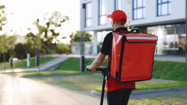 Ένας άγνωστος νεαρός κούριερ καβαλάει το e-scooter του. Παράδοση αρσενικό παράδοση δεμάτων τύπος υπηρεσία βόλτες με ένα ηλεκτρικό σκούτερ κάτω από ένα άδειο πεζοδρόμιο με κόκκινο σακίδιο κάτω από το δρόμο στο ηλιοβασίλεμα — Αρχείο Βίντεο