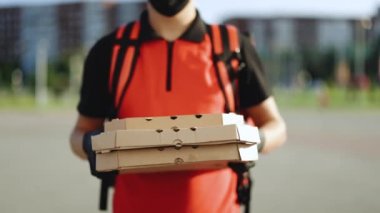 Eldivenli ve tıbbi maske takmış yakışıklı beyaz bir teslimatçının elinde karton kutular tutarken çekilmiş portresi. Erkek kurye pizza veriyor ve kameraya bakıyor.