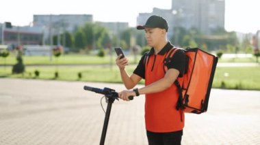 Asyalı kurye yemek teslimatı. Kırmızı termal sırt çantalı. Elektrikli scooterlı caddede yürüyor. Teslimatçı işçi çevrimiçi sipariş müşterisini teslim ediyor.