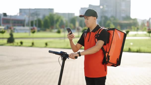 Asijské muž kurýr dodávky potravin s červeným termálním batohem procházky ulicí s elektrickým skútrem používá smartphone navigovat. Dodávka pracovník zaměstnanec doručit on-line objednávky zákazník — Stock video