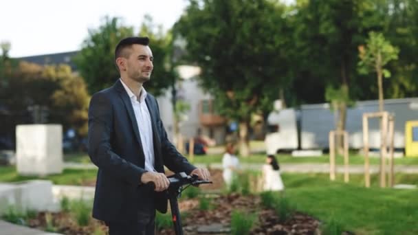 Slow-motion sida-vy av kaukasisk affärsman rider på elektrisk skoter längs trottoaren på varm solig dag. Modern transport gadget och populär futuristisk enhet bland ungdomar. — Stockvideo