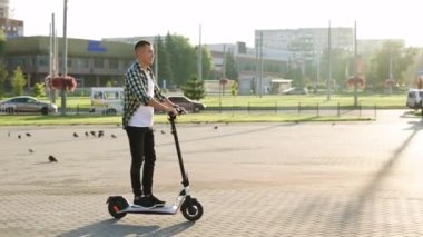 Genç, zeki hippi adam hızlı, modern bir elektrikli scooter kullanıyor. Köy yolundaki açık arazide çağdaş inşaatın yanında. Çevre taşımacılığı. Hızlı sürüş elektrikli taşıma