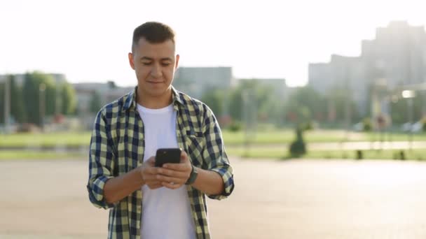 Shot of move around hipster man stand in the city center street uses phone textting scrolling steping smile technology communication sunny day success slow motion. Prohlíží si web na svém zařízení. — Stock video