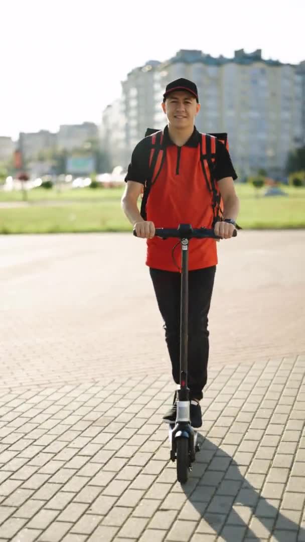 Kırmızı termal sırt çantalı erkek kurye yemek teslimatının dikey videosu elektrikli scooter ile sokakta müşterilere servis yapıyor. Kırmızı üniformalı teslimatçı işçi. — Stok video