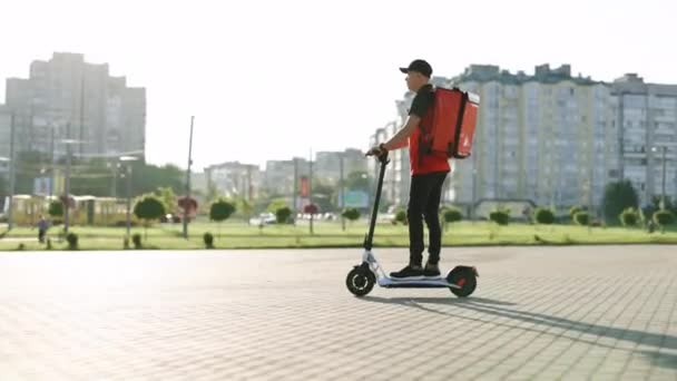 Kurier miejski, kurier w czerwonej koszulce z czerwonym plecakiem termicznym jeździ skuterem elektrycznym. Szybka jazda elektrycznym transportem, poranne światło. Ekologiczny transport — Wideo stockowe