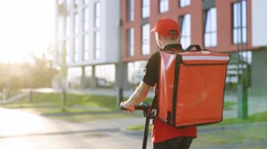 Erkek paket teslimatçısı, gün batımında sokağın aşağısındaki kırmızı sırt çantalı, boş bir kaldırımdan elektrikli scooter kullanıyor. Tanımlanamayan genç kurye e-scooterıyla geziyor.