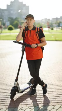 Kırmızı sırt çantalı yemek dağıtım elemanının dikey görüntüsü. Elektrikli scooterlı izotermal yiyecek kutusu olan erkek kurye evin girişine gelir ve müşteri çağırır.