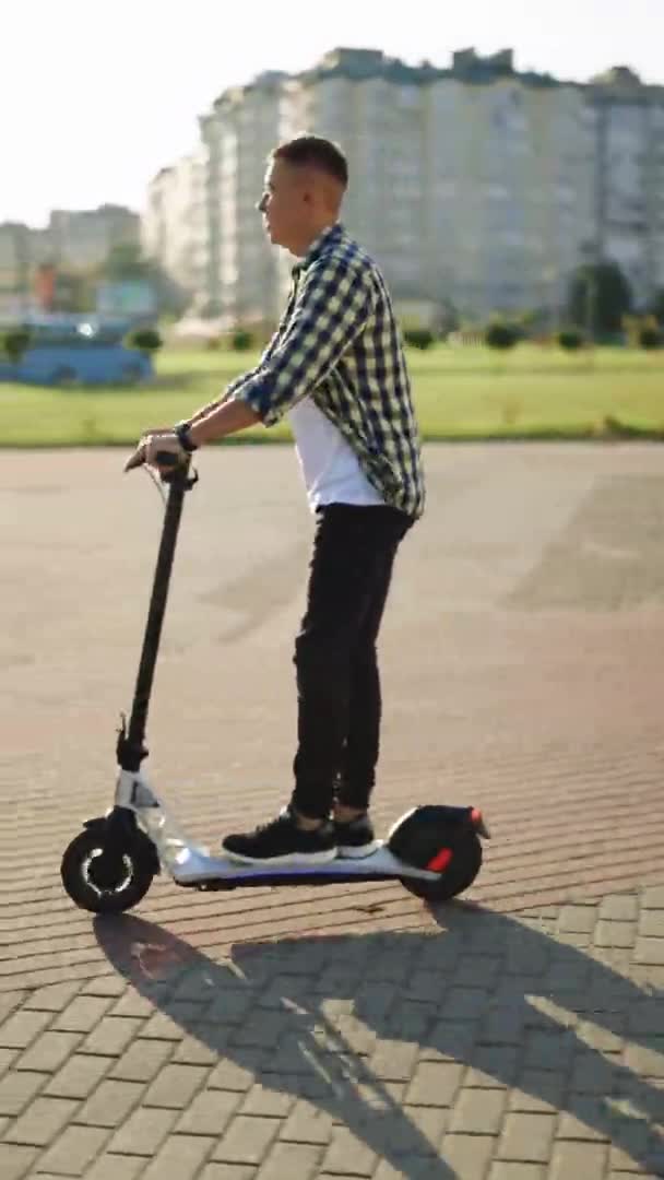 Captura de video vertical de un joven hipster caucásico conduciendo un moderno scooter eléctrico ecológico, transporte moderno en la carretera a la luz de la mañana. Ecología y estilo de vida urbano — Vídeos de Stock