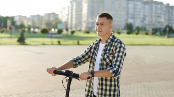 Şehrin önünde elinde e-scooterla duran genç bir adam. Çevre taşımacılığı. Modern kentsel alternatif ulaşım. Çevre dostu ulaşım — Stok video