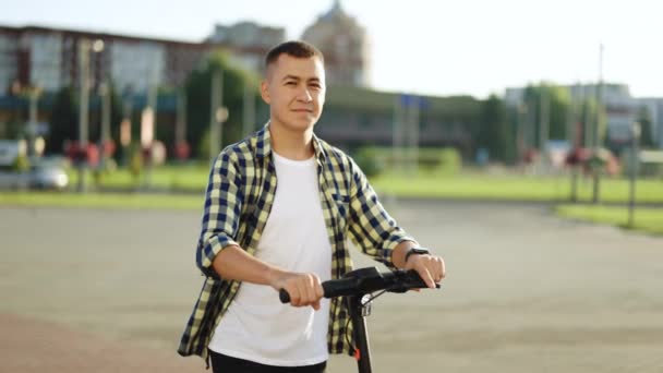 Portraitaufnahme eines kaukasischen Hipster-Mannes, der sich auf einen Elektroroller lehnt und in die Kamera blickt. Stylischer Mann auf Fahrzeug im Freien. Männchen in der Stadt im Sonnenlicht bei Sonnenuntergang. Umweltfreundlicher moderner Nahverkehr — Stockvideo