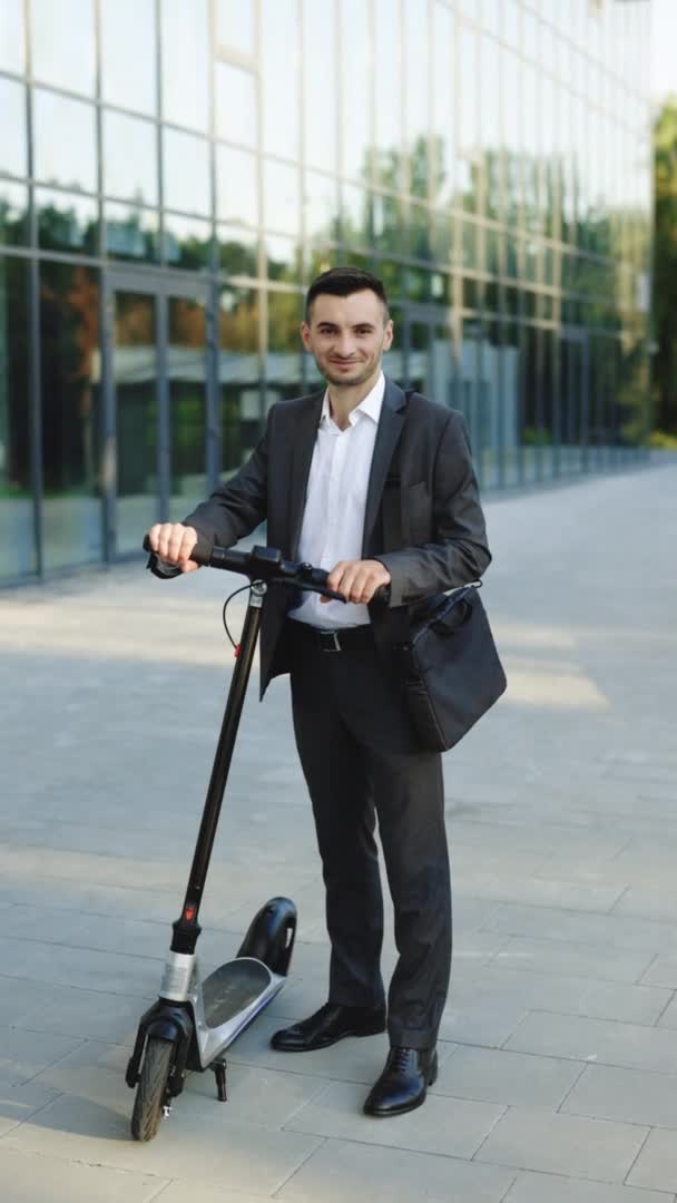 Vídeo vertical de un joven hombre de negocios en traje elegante parado al aire libre, sonriendo alegremente y mostrando el pulgar hacia arriba. Hombre de negocios guapo con su scooter eléctrico en frente del edificio de negocios 4k — Vídeo de stock