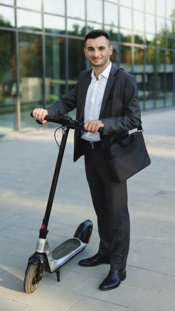 Verticale foto van een zakenman die voor de stad staat, elektrische scooter vasthoudt en naar de camera kijkt. Modern stedelijk alternatief vervoer. Milieuvriendelijk vervoer. Ecologie en stedelijke levensstijl — Stockvideo